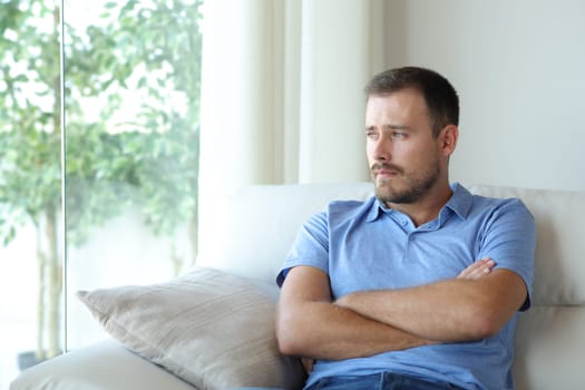 Pensive sad man looking through a window sitting on a couch at home
