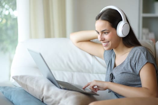 Happy woman wearing headphone watching entertainment content on laptop sitting on a couch at home