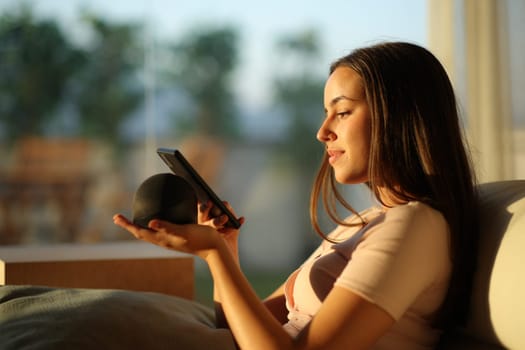 Woman pairing phone with smart speaker at sunset sitting on a couch at home