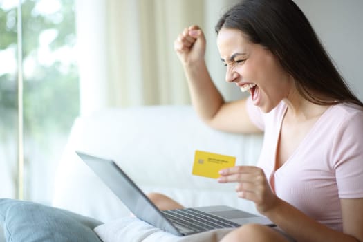 Woman celebrating exciting discount buying online with laptop and credit card sitting on a couch at home