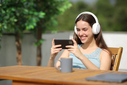 Happy woman wearing headphone in a terrace watching online media on phone a sunny day