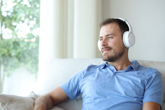 Satisfied man wearing headphone listening audio sitting on a sofa at home looking through a window