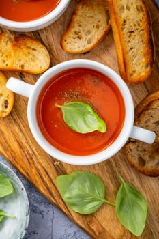 Homemade tomato soup with basil, toast and olive oil on a wooden cutting board.