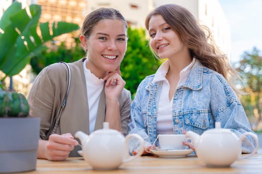 Portrait Two young Caucasian women smiling and looking at the camera while enjoying tea outdoors. Relaxed and cheerful moment of friendship, leisure, and conversation on a sunny day in the city.