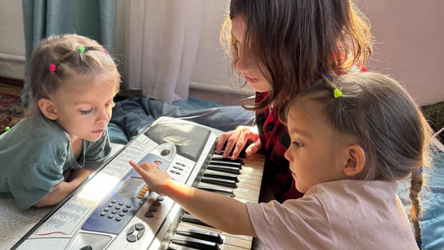 Toddler twin girls and their teenage brother enjoy a musical moment together, exploring sounds on a synthesizer. Fun indoor family learning activity