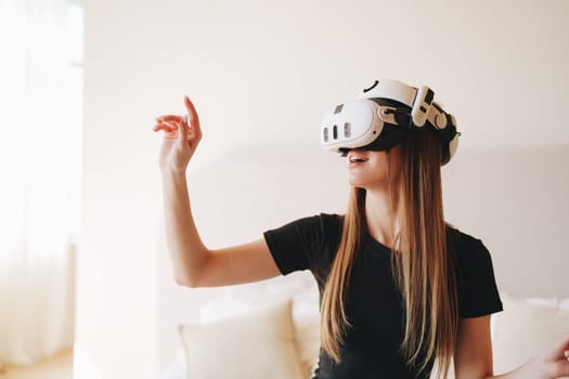 Engaged in a virtual reality experience, a young woman with long hair wears a headset and moves her hand as if reaching out to touch unseen objects while smiling in a well-lit room.