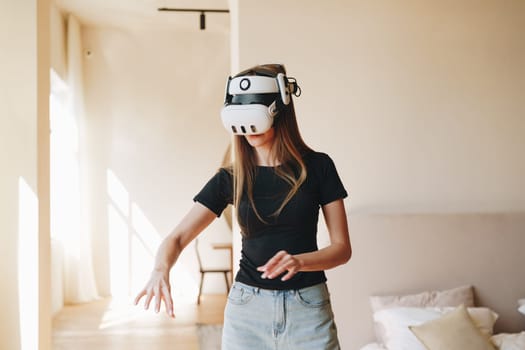 A young woman wearing a virtual reality headset is immersed in an exciting digital world. Sunlight pours through the windows, illuminating her movements and the modern decor.