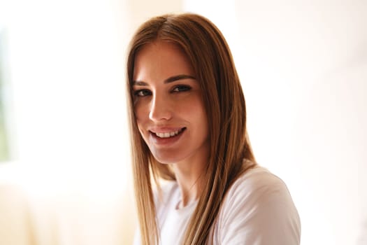 A woman with long, straight hair smiles warmly while sitting in a well-lit indoor space. Sunlight filters through the window, creating a cheerful atmosphere.