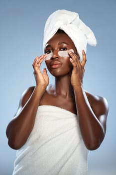A woman is featured preparing for skincare by applying under-eye patches while wrapped in a towel. The calm background highlights her self-care moment.