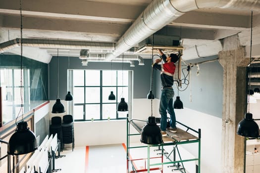 A construction worker is on a scaffold, inspecting and repairing the ceiling equipment in a newly designed office space filled with natural light. Black pendant lights hang overhead.