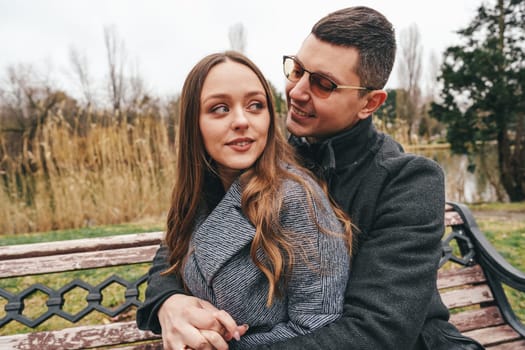A young couple sits closely on a bench in the park, sharing smiles and laughter as they enjoy a cool, overcast day amid nature's beauty and tranquility.