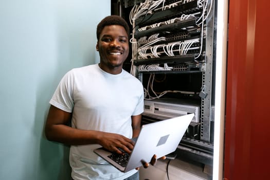 A young professional stands beside server racks, smiling widely while using a laptop. The environment is a data center, highlighted by multiple cables and equipment.