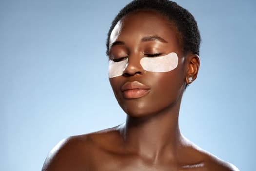 A woman is sitting peacefully with her eyes closed, applying under-eye patches to revitalize her skin. The serene atmosphere enhances her skincare routine in a studio setting.