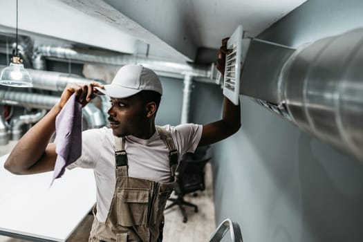 A maintenance worker in a stylish office adjusts an air vent while holding a cloth to remove dust. The setting features metal ducts and a contemporary workspace.