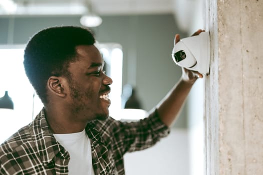 A young man with short hair smiles while installing a security camera on a wall in a contemporary office setting. Bright lighting enhances the warm atmosphere.