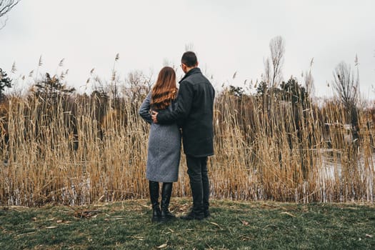 A couple stands close together, gazing at the water while surrounded by tall grass. The day is cloudy, creating a serene atmosphere for their intimate moment.