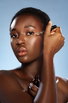 A woman with short, natural hair is delicately applying a serum to her cheek using a dropper. She showcases a glowing complexion in a well-lit studio with a blue background.