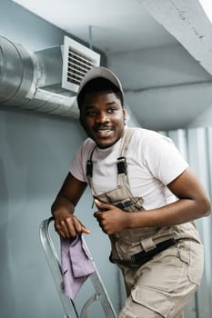 A young worker is on a ladder, smiling and holding a purple cloth. He is dressed in beige overalls and a cap, ready to perform maintenance in a well-lit indoor area.