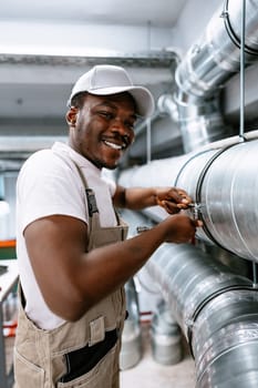 A man in a cap smiles while using a wrench to secure ductwork in an industrial setting. Bright lighting emphasizes his focused expression and the well-maintained environment.