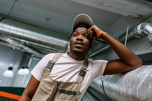 A young man wearing a light cap and beige overalls leans against a metallic surface in an industrial building. He appears deep in thought, contemplating his day.