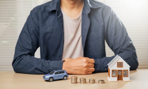 Man sitting with small car and house models beside coin stacks on a table. Concept of finance, saving for property, auto loans, down payments, installment costs, and tax strategies for homeownership.