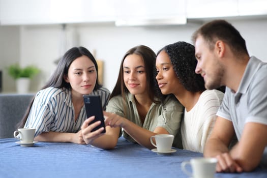 Interracial friends checking mobile phone content together in a kitchen at home