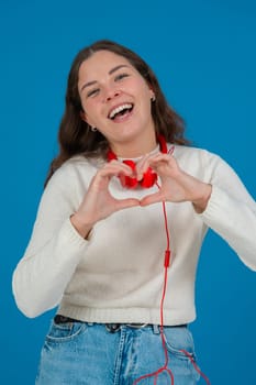 Happy young woman in white sweater and red headphones smiling while making a heart shape with her hands against a blue background, representing kindness, connection, love, and positive emotions. Vertical