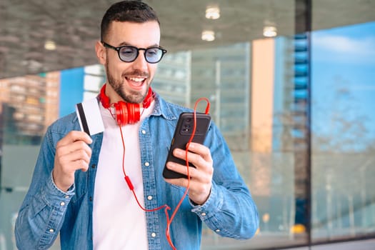 Happy man celebrating a successful online purchase while holding a credit card and smartphone. He smiles with excitement, wearing headphones and sunglasses in a modern urban environment.