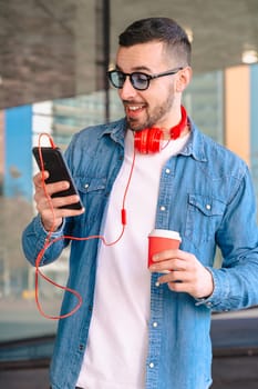 Young man with red headphones enjoying coffee while using his smartphone in a city environment. He smiles while reading or texting, standing outside modern glass buildings in a casual moment. Vertical
