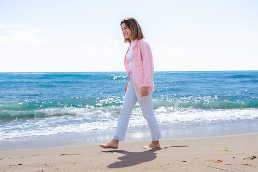Happy woman walking barefoot on the beach, enjoying a peaceful sunny day by the sea. She wears casual clothes and a smile, connecting with nature in a calm and refreshing coastal environment.