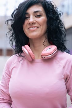 Individual portrait of a Caucasian woman wearing a pink hoodie and headphones outdoors. She smiles slightly, standing confidently in an urban environment with a casual and relaxed look. Vertical