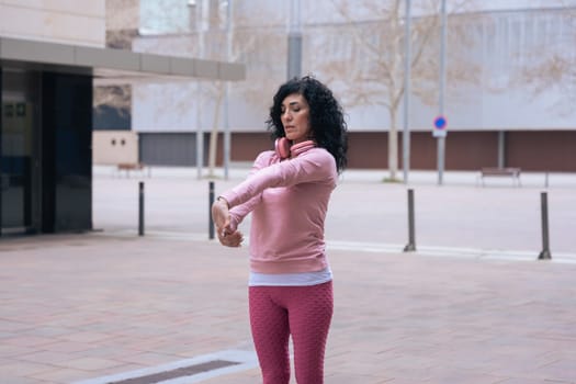 Caucasian adult woman with curly black hair stretching her arms outdoors in an urban setting.she is wearing a pink sweatshirt and exercising for a healthy lifestyle, fitness, and wellness in the city