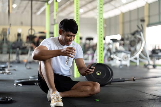 A young man sits on the gym floor, enjoying a moment of relaxation while checking his phone and sipping water after an intense workout.