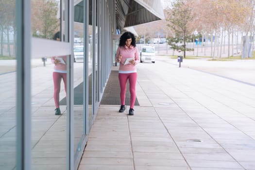Confident caucasian woman in pink activewear holding documents and standing near a glass building. Modern urban lifestyle, fitness, and casual fashion concept in a contemporary outdoor environment.
