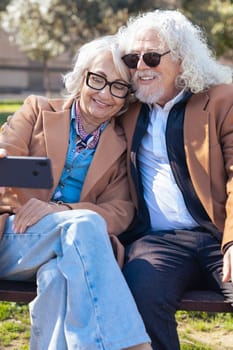 Elderly couple dressed in brown coats taking a cheerful selfie outdoors. They smile while holding a smartphone, capturing a happy moment together in a sunny and casual environment.Vertical