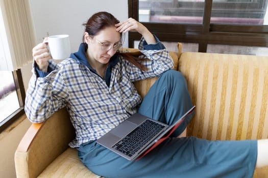 Woman sitting comfortably on sofa, working on laptop while savoring a cup of coffee in a moment of relaxation