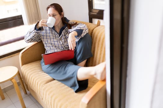 Woman enjoying a cup of coffee while relaxing on her sofa with a tablet, working from home