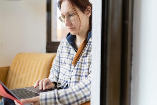 Focused female freelancer working remotely using laptop and consulting a book, sitting comfortably on a sofa