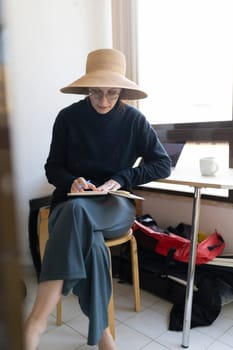 Woman wearing straw hat taking notes on notebook sitting on stool at home