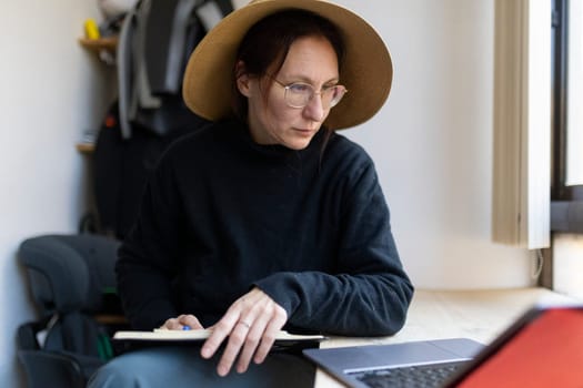 Woman working from home using laptop and taking notes, wearing straw hat and glasses