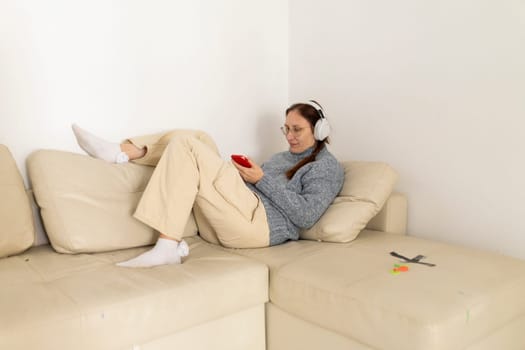 Woman wearing headphones is lying on a sofa, using smartphone and listening to music