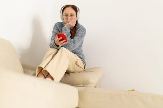 Bored mature woman wearing wireless headphones and browsing her smartphone while sitting on a sofa at home