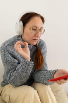 Redhead woman wearing headphones and holding smartphone is making ok sign with fingers