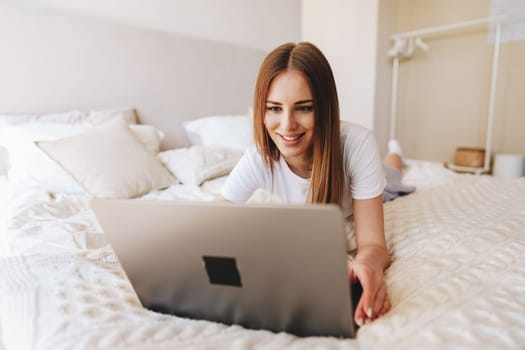 A woman with long brown hair smiles while using a laptop on her bed. Soft pillows and a warm ambiance create a comfortable and inviting atmosphere in her personal space.
