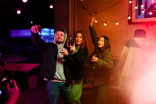 Three friends celebrate together outside, holding glasses of wine and posing cheerfully under string lights. The festive ambiance enhances their night of laughter and joy.