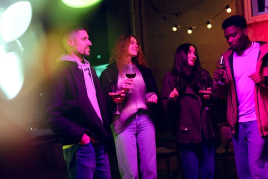 A group of four friends is gathered outdoors, holding wine glasses and chatting animatedly. String lights illuminate the atmosphere, enhancing the festive vibe of the evening.
