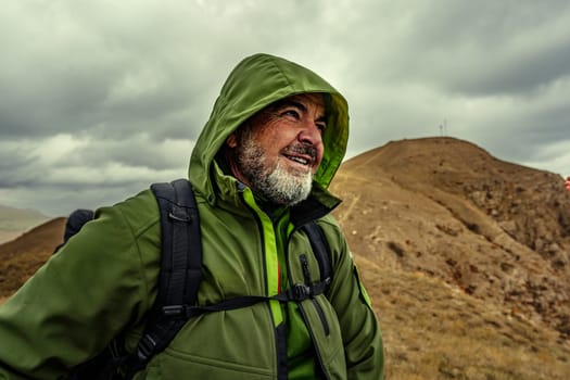 A determined man wearing a green jacket smiles as he navigates the hilly landscape under a moody sky, showcasing a spirit of adventure and exploration in nature.