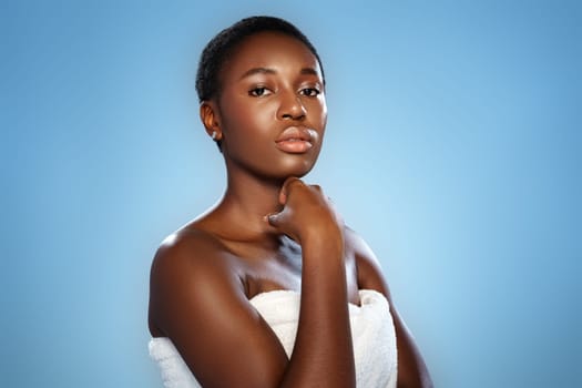 A model stands in a graceful pose, highlighting her features with a soft expression. The blue backdrop enhances her natural beauty, creating a serene atmosphere.