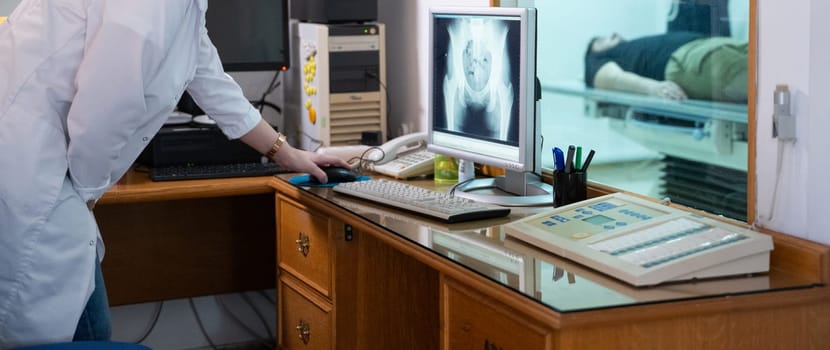 A radiologist in a white lab coat sitting at a workstation while analyzing an X-ray image of a patients hip in a medical imaging room.