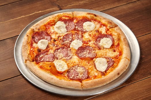 Round pizza with tomato, salami, cream cheese and grated cheese on metallic tray over wooden table